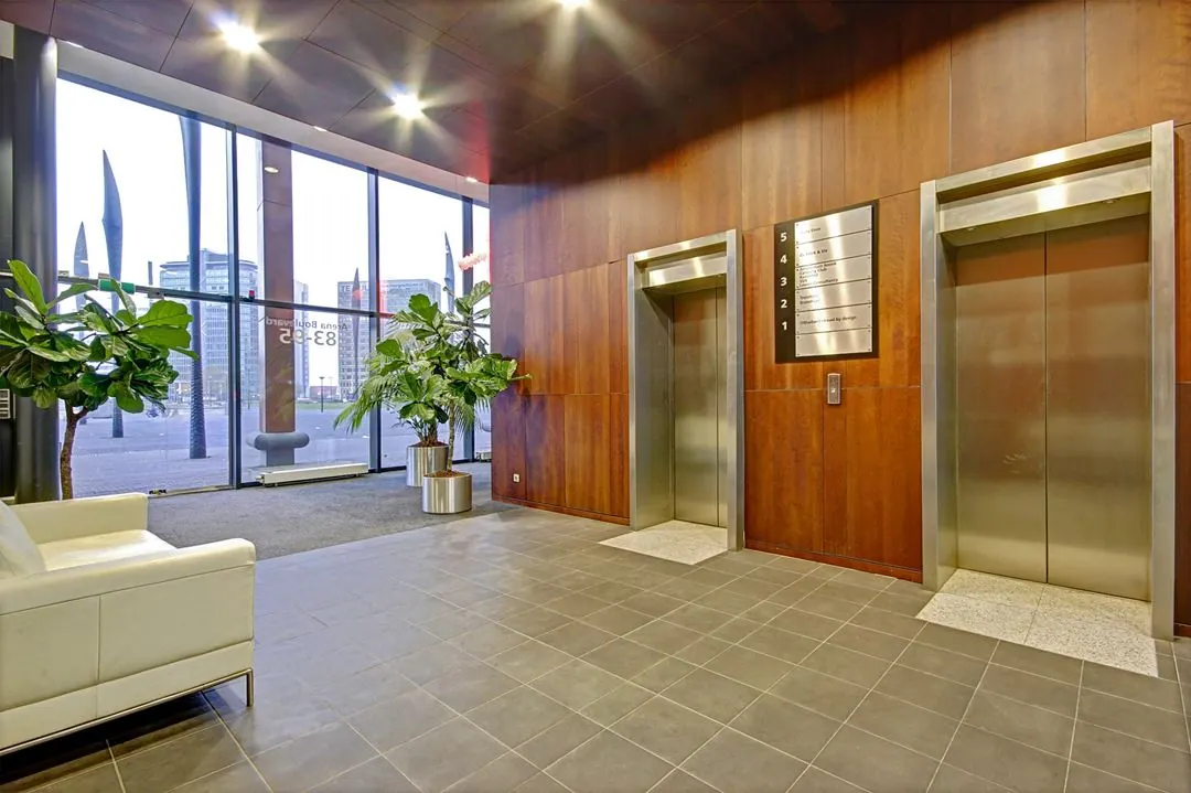 Moderne entreehal aan de Johan Cruijff Boulevard met liften, grote kamerplanten en panoramisch uitzicht op kantoorgebouwen.